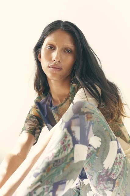 Portrait of a young woman in patterned top and pants, seated with legs crossed against a light background, styled with facial piercings.