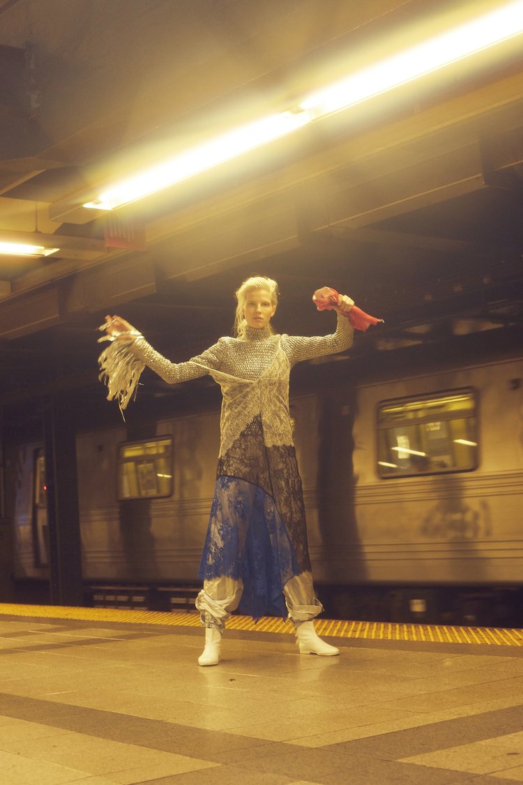 Fashion photography of a woman in an avant-garde outfit in a subway station, featuring a train, platform, and yellow lighting.