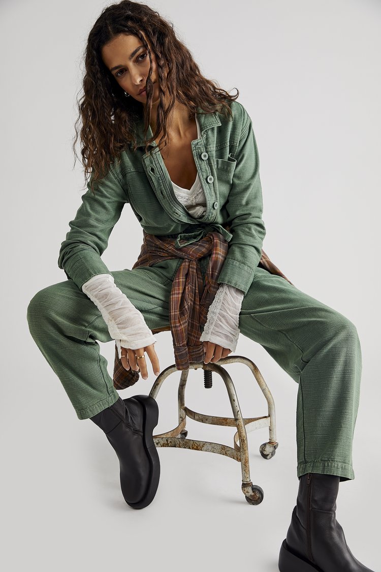 A woman with long brown hair wearing a sage green jumpsuit and black boots sits on a metal stool in a brightly lit studio setting.
