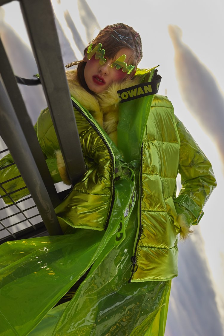 Young woman with artistic visor and lime green puffy jacket posing in front of structural background with fair lighting and high fashion appeal.