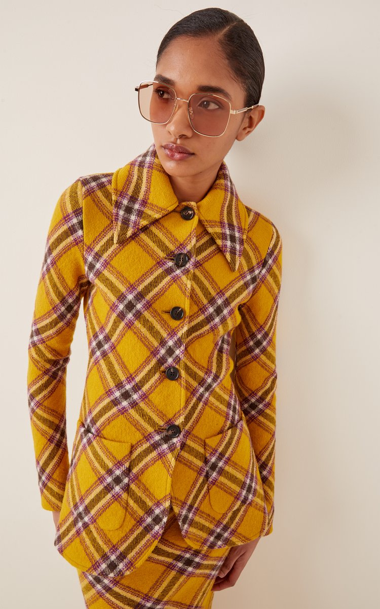 Portrait of an adult woman with tan skin wearing a yellow plaid jacket and sunglasses, posing in a brightly lit, neutral studio.
