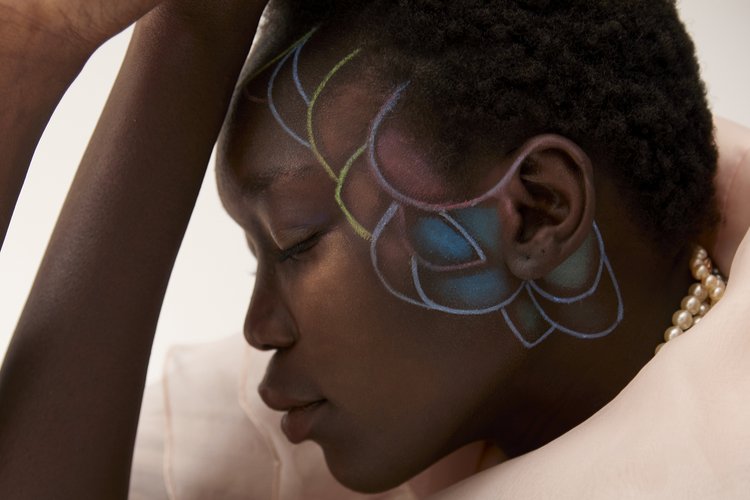 Close-up portrait of a Black woman with colorful chalk drawing on her face and neck, wearing a pearl necklace.
