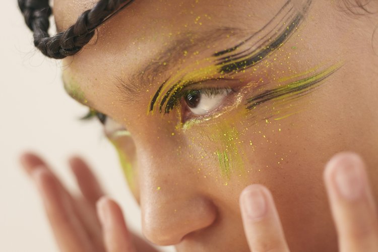 Close-up of an adult female face with yellow and black eyeshadow makeup, light skin tone, and braided hair against a neutral background.