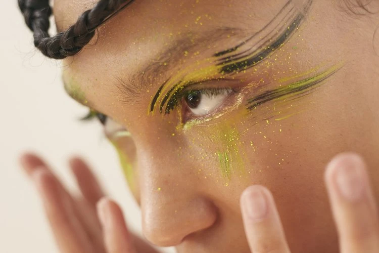 Close-up of an adult female face with yellow and black eyeshadow makeup, light skin tone, and braided hair against a neutral background.