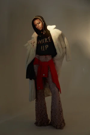 Full-length fashion shot of a black female in a hooded sweatshirt with lace pants and a white coat in a studio with a warm lighting.