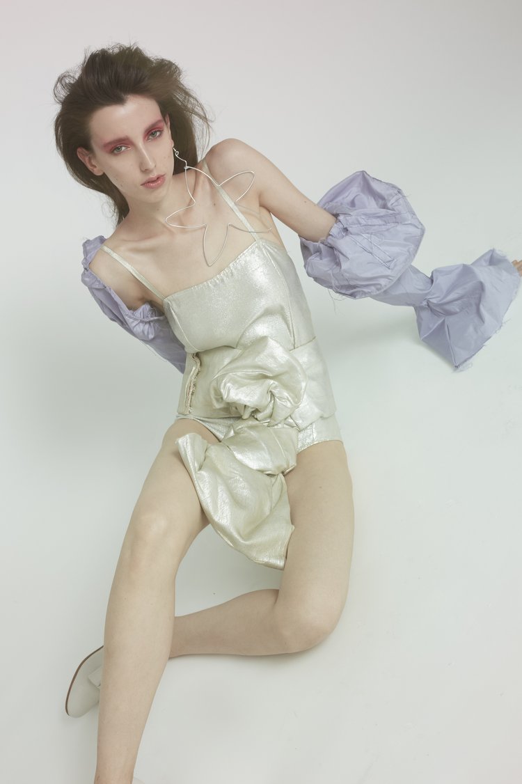 An adult female model in a futuristic silver bodysuit with dramatic makeup poses in a studio setting with a seamless white background.