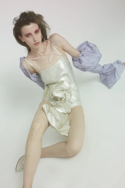 An adult female model in a futuristic silver bodysuit with dramatic makeup poses in a studio setting with a seamless white background.