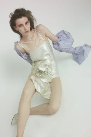 An adult female model in a futuristic silver bodysuit with dramatic makeup poses in a studio setting with a seamless white background.