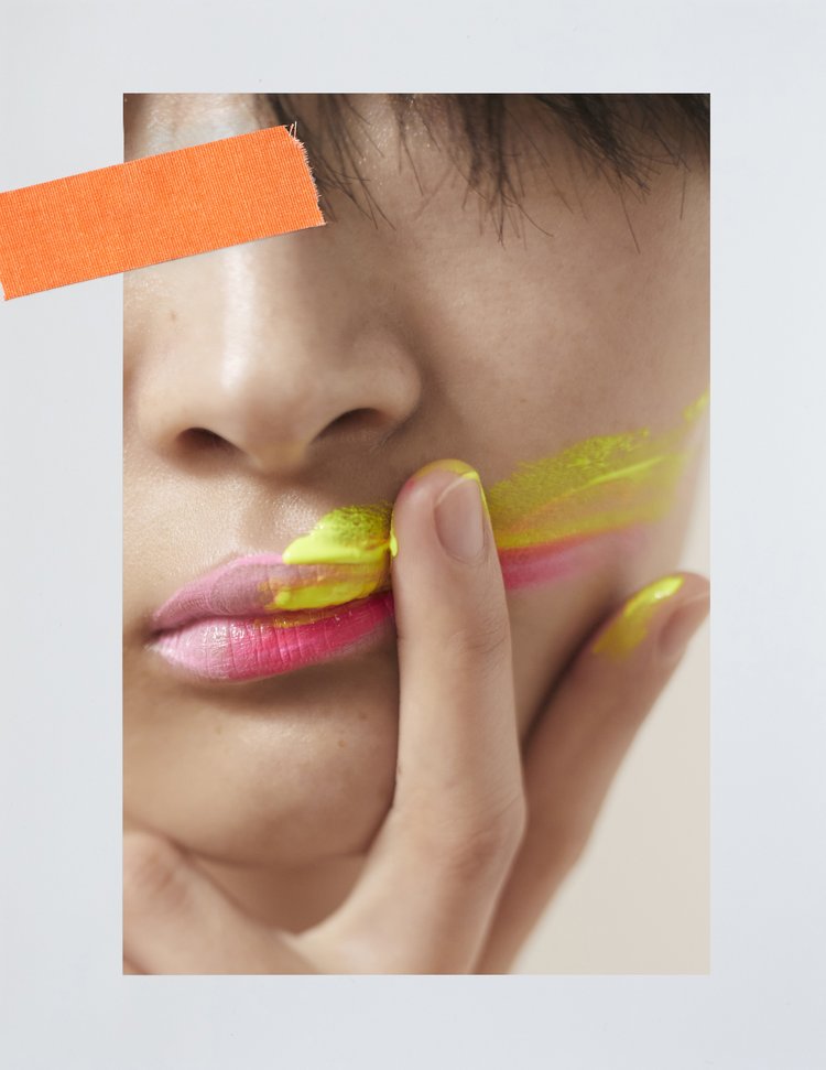 Cropped portrait of a woman with pink and yellow paint on her lips and face, with an orange strip of tape in the background.