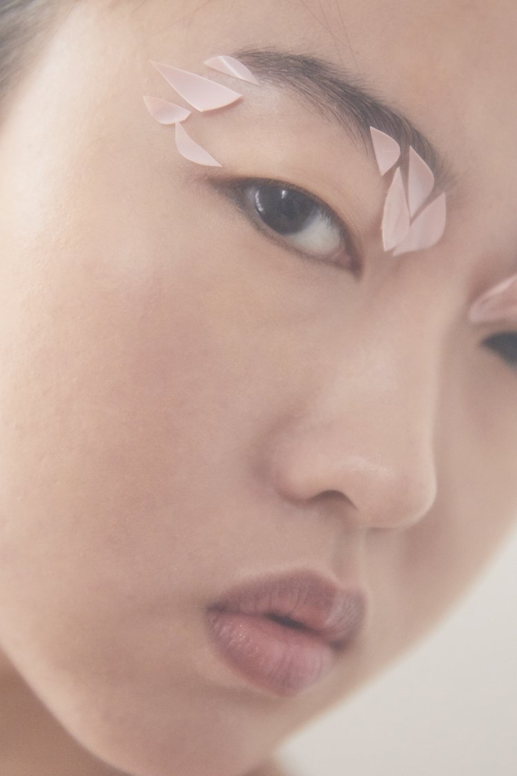 Close-up portrait of a woman with light skin and light pink feathers on her eyebrows, against a neutral backdrop, beauty.