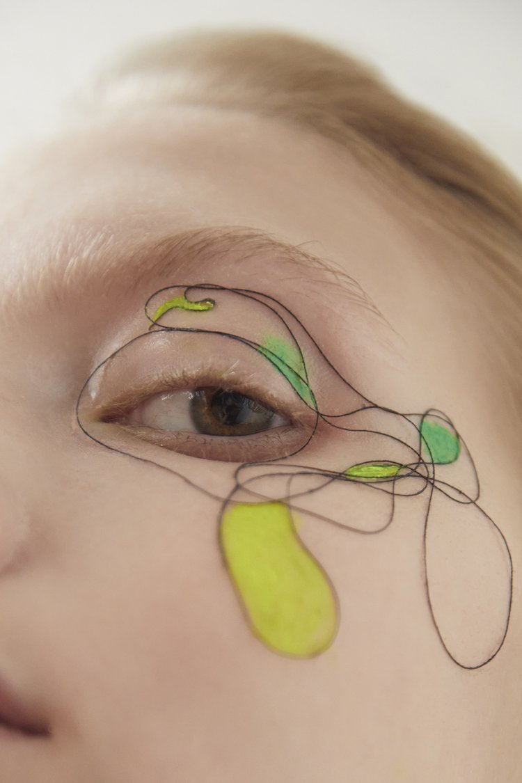 Close-up of a woman's face featuring makeup with freehand lines and shapes in shades of black, green, and yellow around the eye.