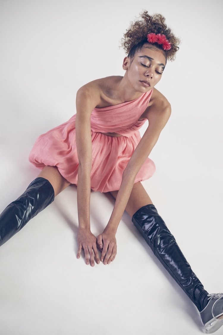 Woman in a pink dress and thigh-high boots sits on a white background. Portrait with eyes closed and a floral headband.