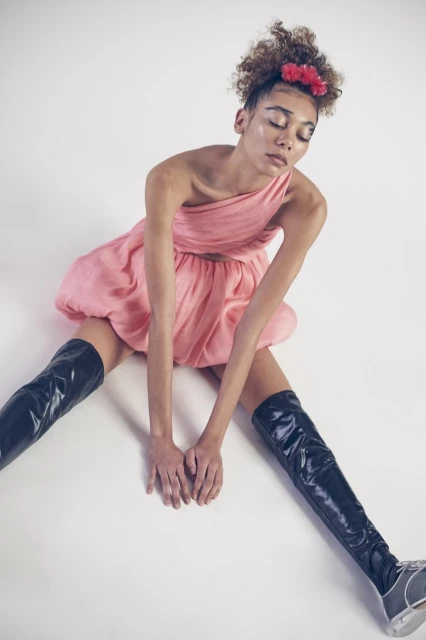 Woman in a pink dress and thigh-high boots sits on a white background. Portrait with eyes closed and a floral headband.