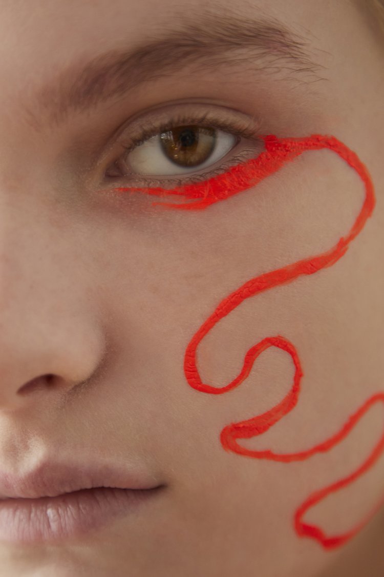 Close-up portrait of a fair-skinned person with striking neon orange graphic eyeliner design extending onto the cheek, focusing on eye detail.