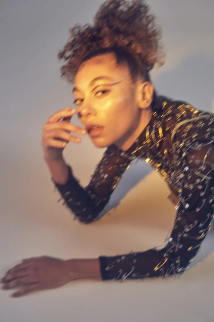 Close-up of a young adult female model posing in a translucent, black, long-sleeved top decorated with silver safety pins, against a white background.