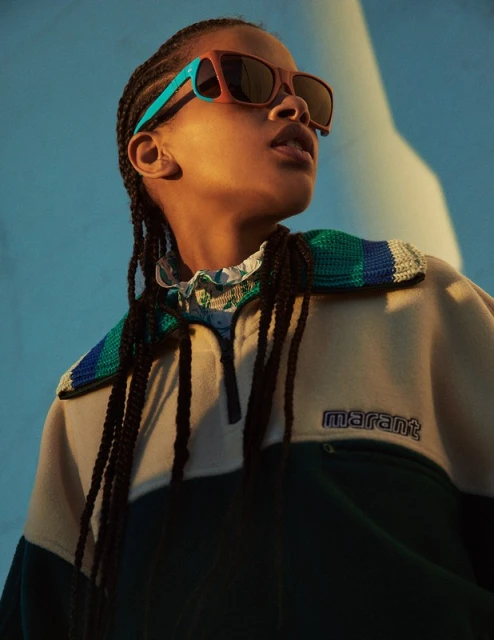 Woman with braided hair poses in a studio wearing sunglasses and a color-blocked shirt with the brand name 'Marant' visible.