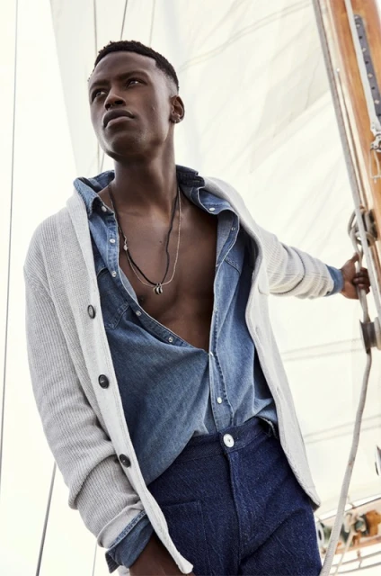 Male fashion model poses in an unbuttoned denim shirt and cardigan on a sailing vessel with white sail in the background.