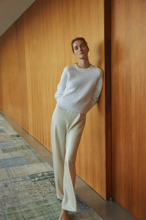 A slender woman with her hair up leans against a wood paneled wall wearing a white sweater and tan slacks in a fashion style photograph.