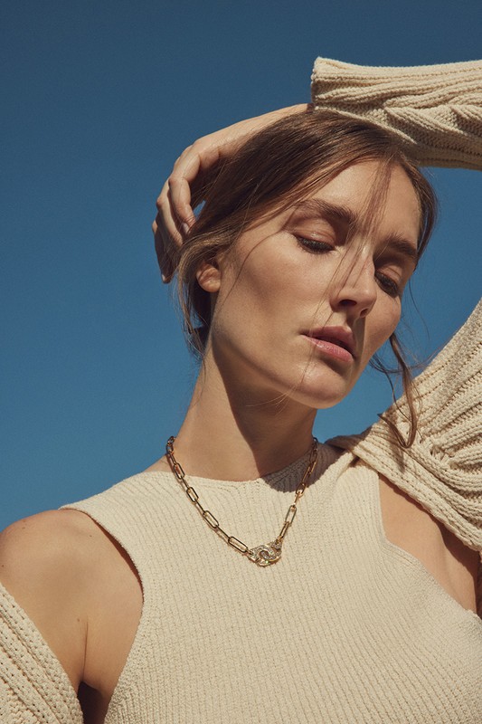 Woman with closed eyes poses in front of a bright blue sky wearing a gold chain necklace, a white tank top, and a cream-colored sweater.