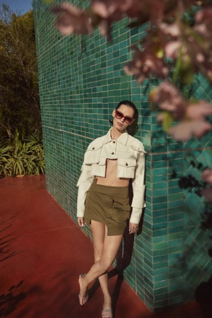 A woman in sunglasses poses against a turquoise tiled wall in a white crop top and short olive skirt in an outdoor setting.