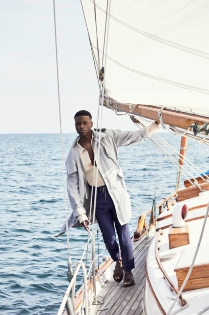 Fashion shot of an adult male on a sailboat, wearing a trench coat, pants, and sandals, posing on deck with the ocean and sky visible.