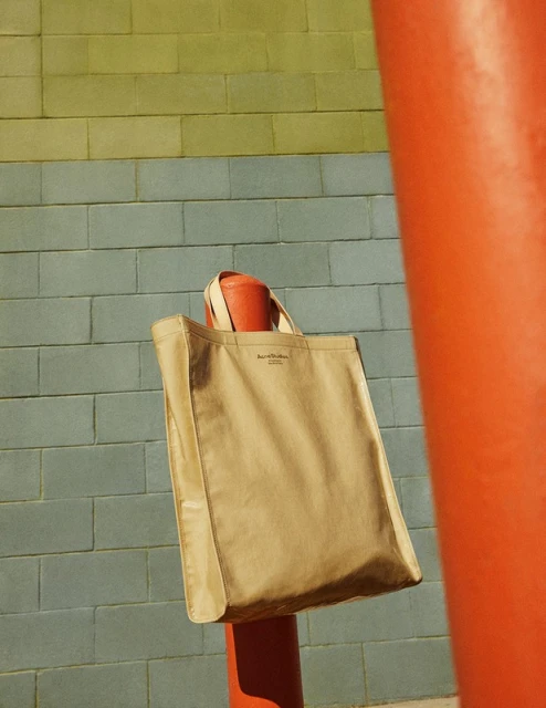 Tan canvas tote bag with leather straps, hanging on a red painted metal post in front of a neutral-colored brick wall.