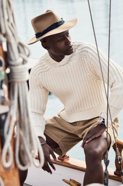 Stylish man with hat and cream sweater on wooden boat with calm water backdrop, outdoor fashion shot, demonstrating summer nautical style.