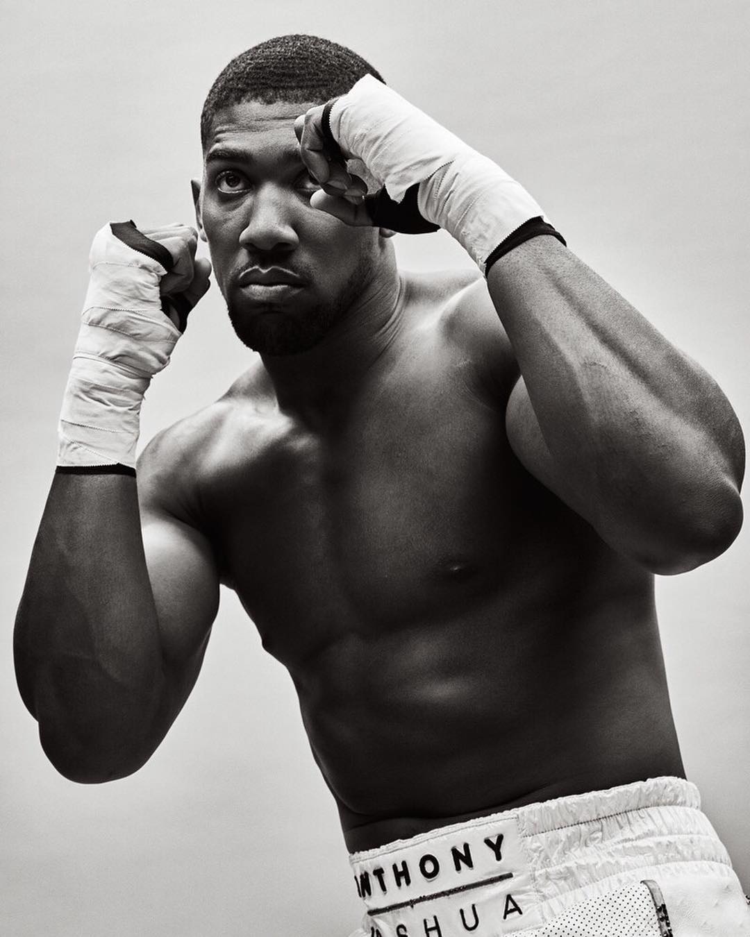 Monochrome portrait of muscular boxer Anthony Joshua in fighting stance, bare-chested with wrapped hands, focused expression, studio lighting.
