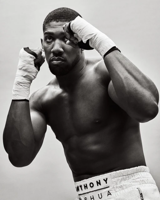 Monochrome portrait of muscular boxer Anthony Joshua in fighting stance, bare-chested with wrapped hands, focused expression, studio lighting.