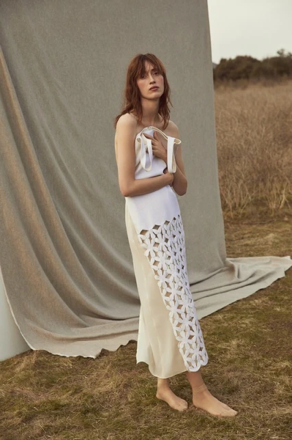 Elegant woman in a white cut-out dress poses outdoors, blending fashion with nature against a beige backdrop and grassy field.