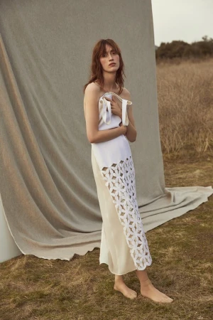 Elegant woman in a white cut-out dress poses outdoors, blending fashion with nature against a beige backdrop and grassy field.