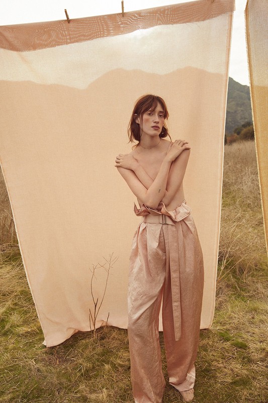 A woman with fair skin poses in wide, light pink pants against an outdoor background with a peach-colored sheet hanging on a line.