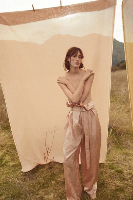 A woman with fair skin poses in wide, light pink pants against an outdoor background with a peach-colored sheet hanging on a line.