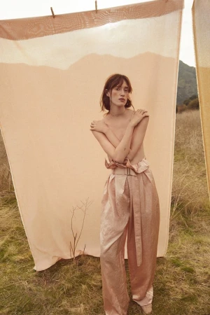 A woman with fair skin poses in wide, light pink pants against an outdoor background with a peach-colored sheet hanging on a line.