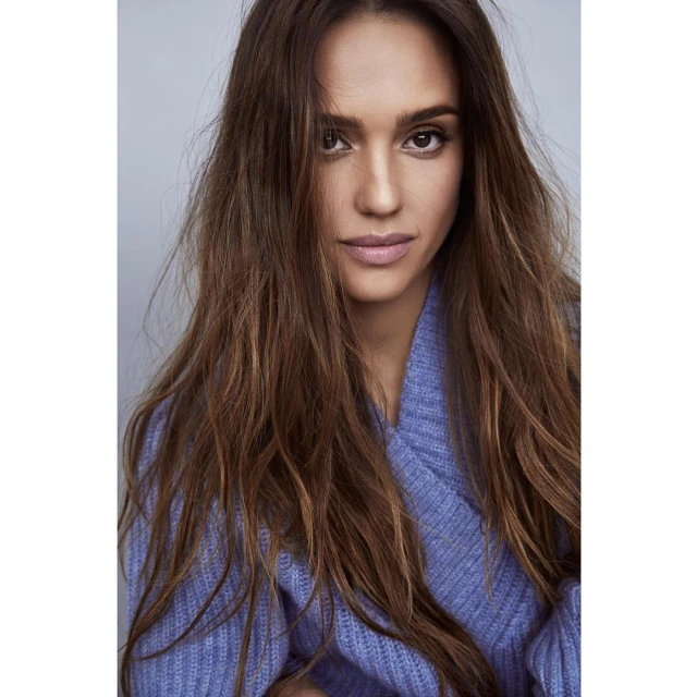 Close-up portrait of Jessica Alba with long brown hair wearing a blue sweater against a neutral grey background in a photo studio.