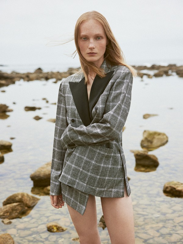 A woman with red hair wearing a gray plaid blazer and skirt poses on a rocky beach with the ocean in the background, shot on an overcast day.