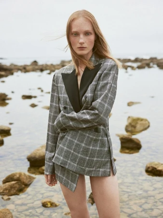 A woman with red hair wearing a gray plaid blazer and skirt poses on a rocky beach with the ocean in the background, shot on an overcast day.
