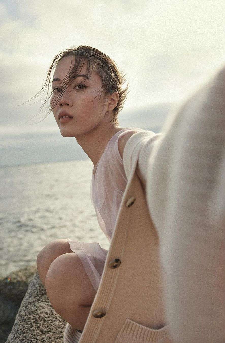 Woman in pink dress and cardigan crouching on rocky coast by the ocean, captured by Williams + Hirakawa.