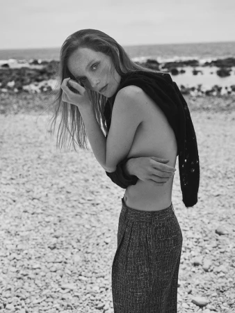 Model with long hair standing on a rocky beach wearing patterned trousers and a dark sweater draped over her shoulders