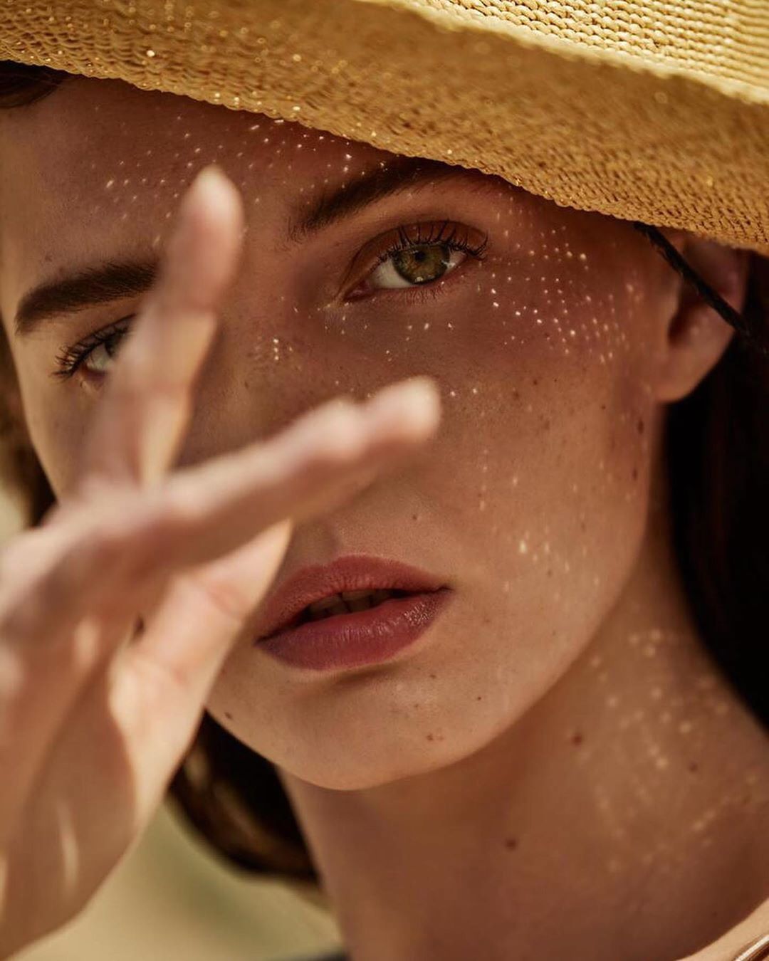 Close-up of a woman with freckles wearing a straw hat, partially obscuring her face with her hand in a bright, outdoor setting.