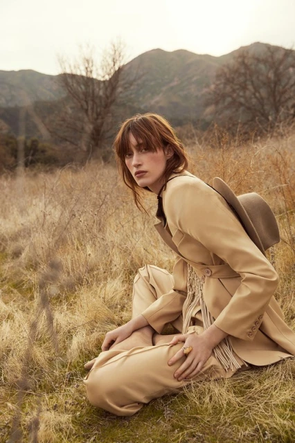 Adult female model poses in a beige pantsuit and hat, seated in a grassy field with distant mountains under soft daylight.