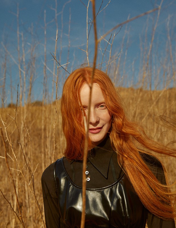 Portrait of an adult woman with red hair and a black top posing in a dry grass field under a blue sky on a sunny day.