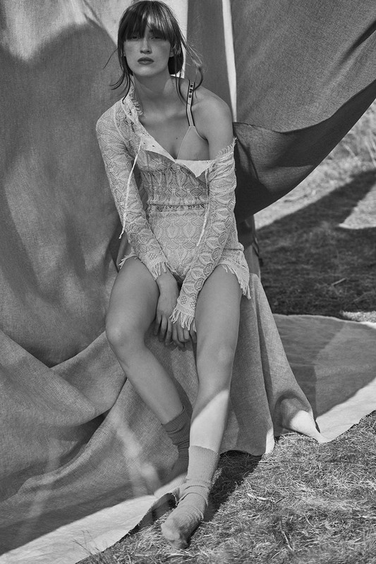 A person sits draped with fabric in a sunlit outdoor setting, wearing a lace outfit and socks, rendered in black and white.