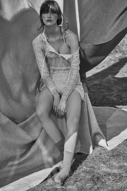 A person sits draped with fabric in a sunlit outdoor setting, wearing a lace outfit and socks, rendered in black and white.