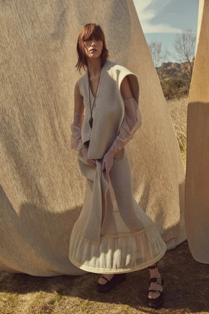 Fashionable woman posing in neutral-toned clothing with gloves in outdoor setting with dry grass and large canvas-like backdrop.
