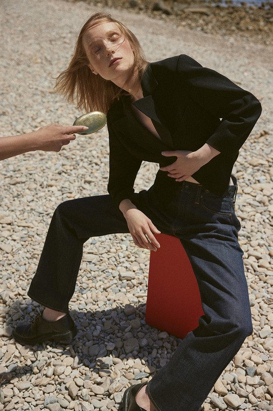 Fashion editorial shot of a woman in dark blazer and jeans sitting outdoors on a red prop, another person holding a makeup compact.