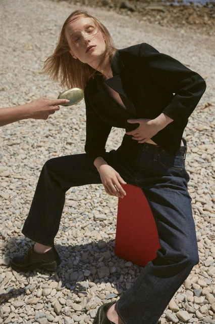 Fashion editorial shot of a woman in dark blazer and jeans sitting outdoors on a red prop, another person holding a makeup compact.