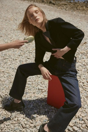 Fashion editorial shot of a woman in dark blazer and jeans sitting outdoors on a red prop, another person holding a makeup compact.
