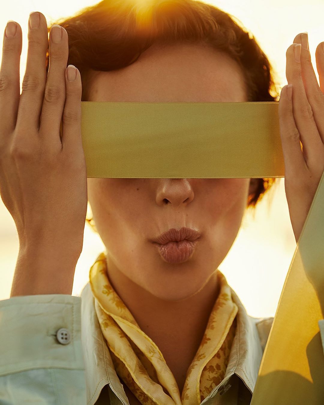 Close-up shot of a young adult woman with a yellow scarf, holding a yellow paper rectangle in front of her eyes, lit by golden light.