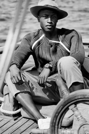 Black man wearing a fedora hat and ribbed sweater seated on a wooden boat deck photographed by Williams + Hirakawa.